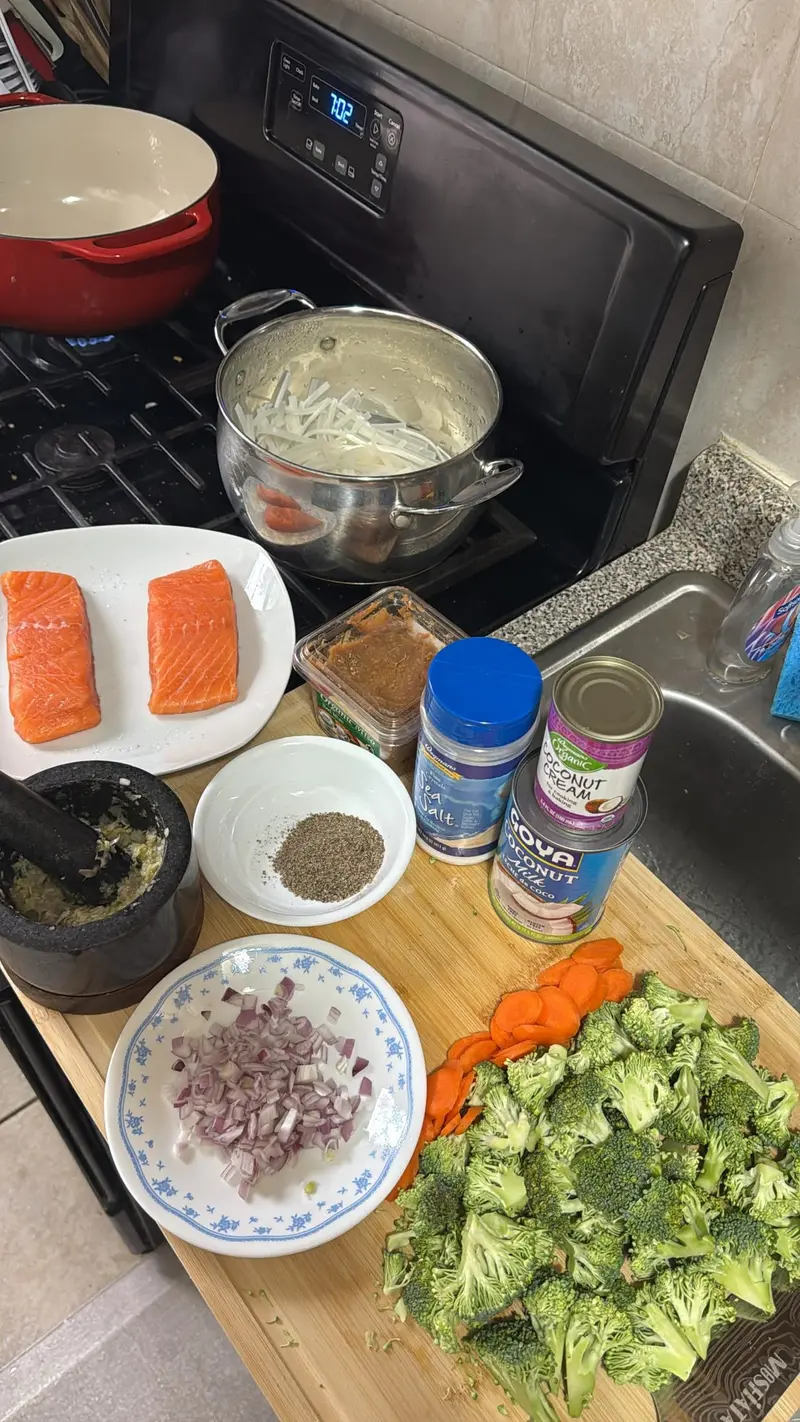 Salmon, broccoli, carrots, red onion, coconut milk, seasonings, and noodles arranged beside pots on a stovetop.