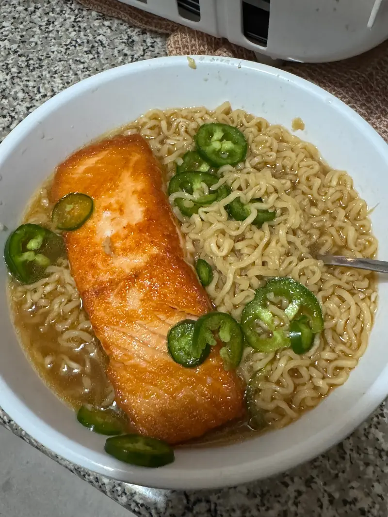 A browned salmon fillet in broth with noodles and sliced green chilis in a white bowl.