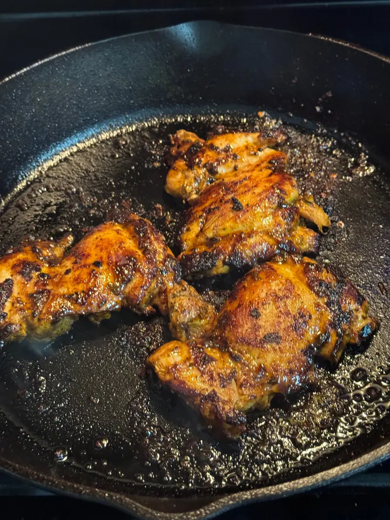 Several deeply browned pieces of seasoned chicken cooking in a cast-iron skillet.