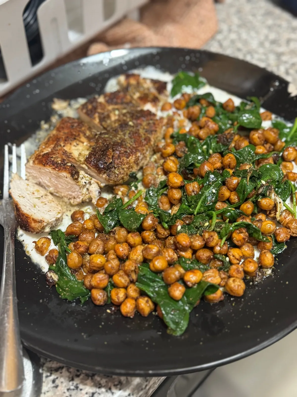 Sliced roasted meat with crisp chickpeas and wilted spinach over a white sauce on a black plate.