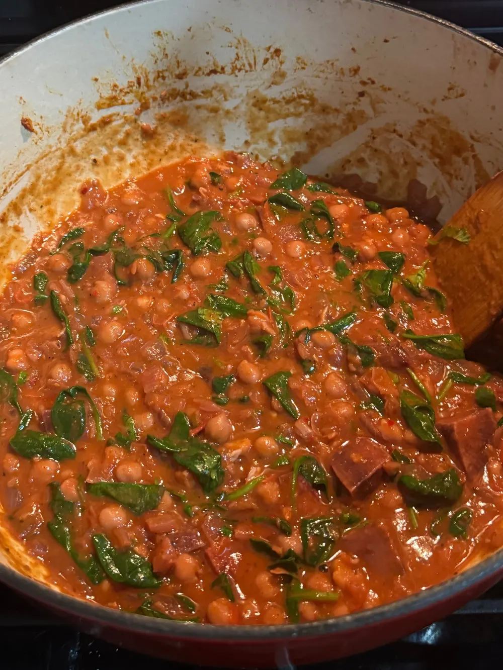 Chickpeas and chopped spinach simmering in a thick red-orange sauce inside a white Dutch oven.