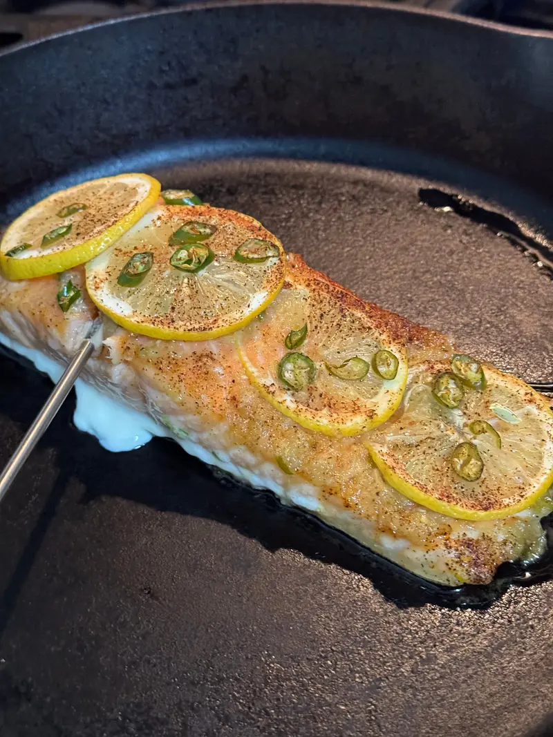 A fish fillet cooking in a cast-iron skillet beneath lemon slices, green chilis, and black pepper.