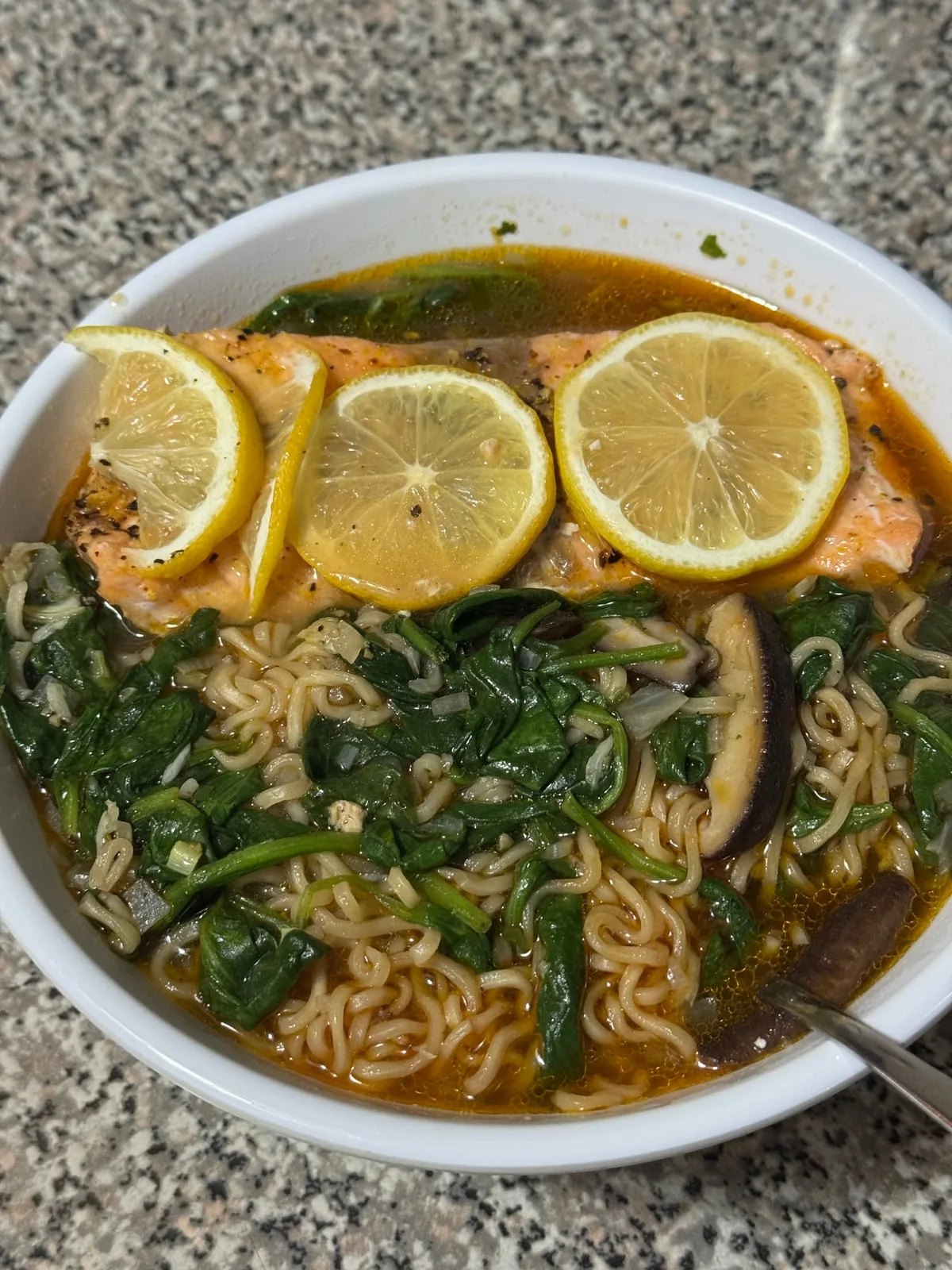 Salmon topped with lemon slices in a bowl of noodles, spinach, mushrooms, and golden broth.